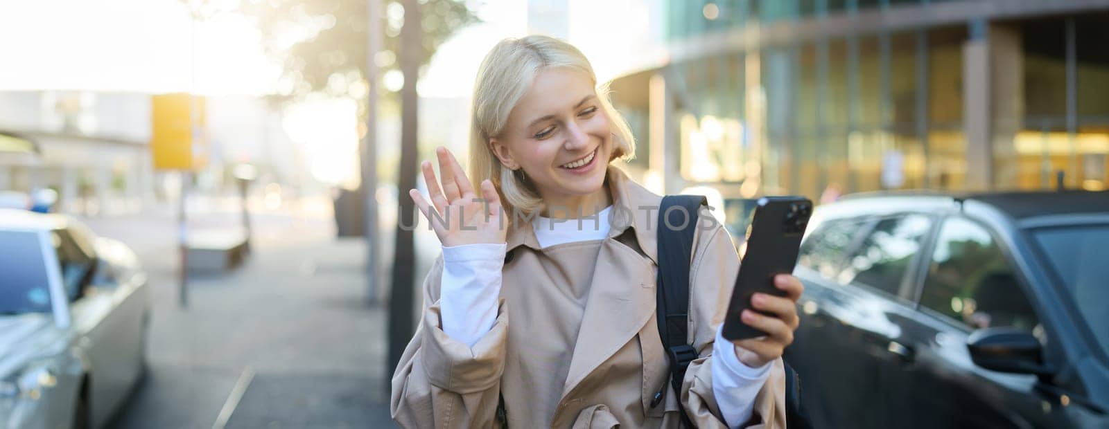 Smiling young modern woman, walking on street, video chats with friend, says hello, waves hand at mobile phone camera, talking to someone on smartphone application.