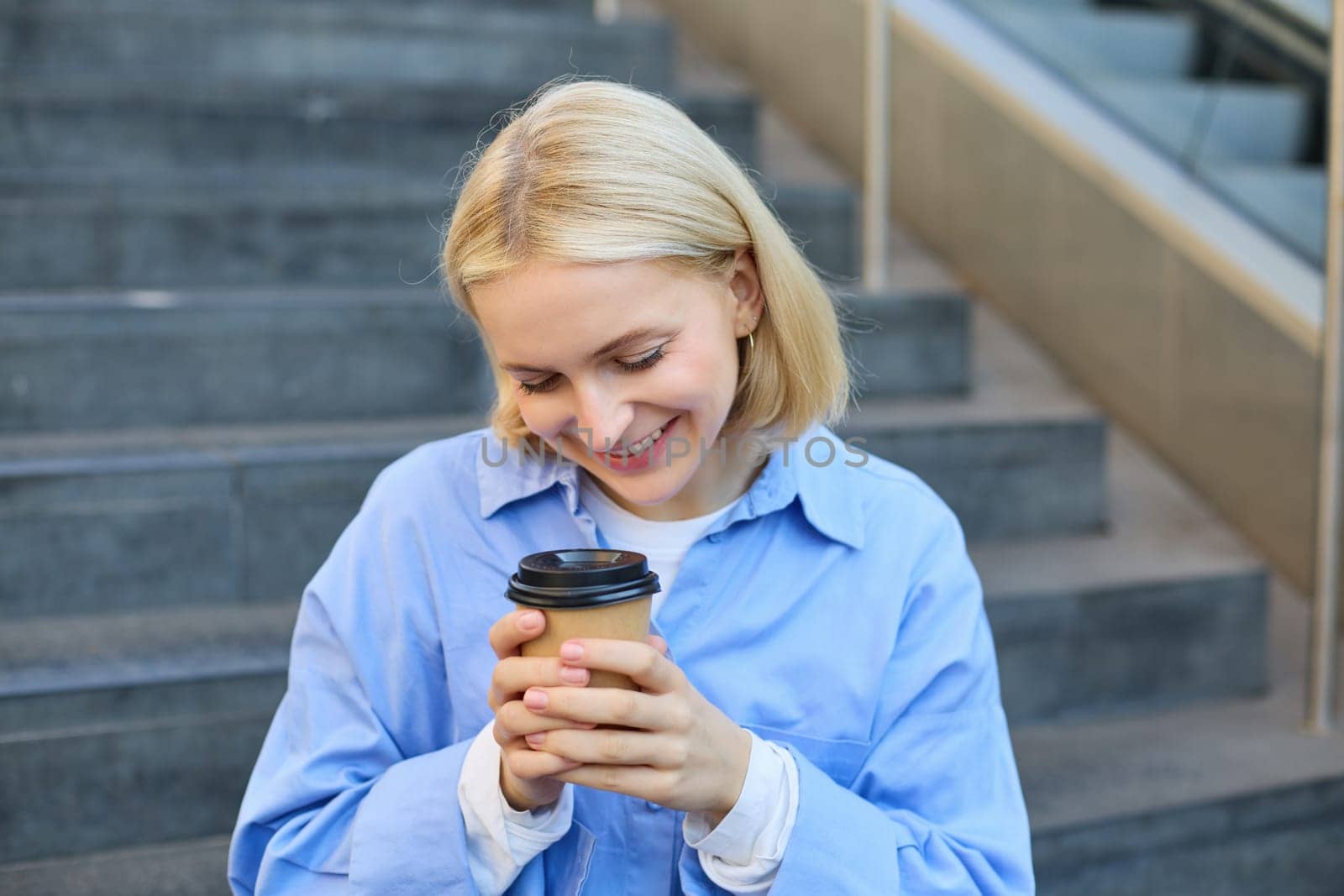 Cute young female student, woman with cup of coffee in hands, resting on stairs in city centre, sitting and drinking cappuccino, smiling happily by Benzoix