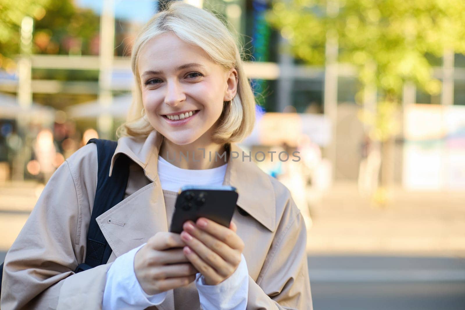 Close up portrait of smiling, beautiful blond girl, holding backpack and mobile phone, standing on street with lots of sunlight, buildings and road behind, looking happy at camera by Benzoix