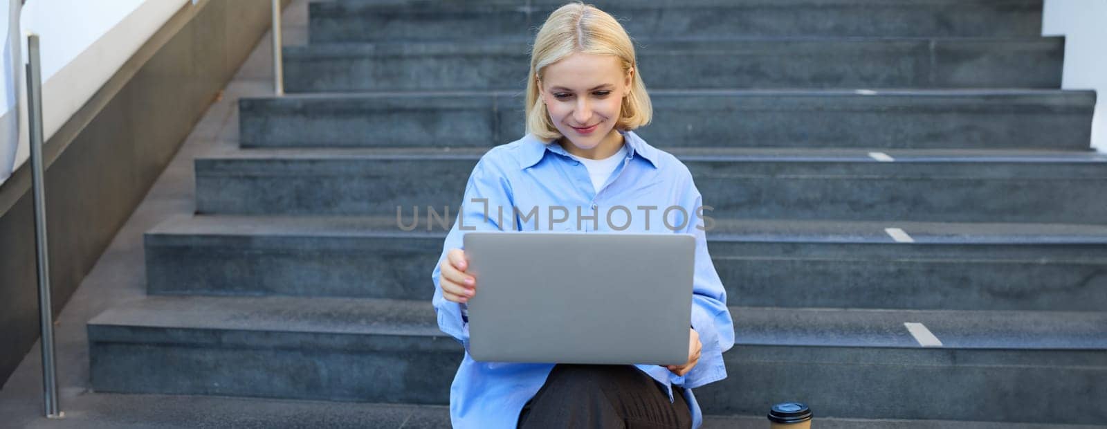 Lifestyle shot of young woman sitting on stairs with laptop, studying outdoors, drinking cup of coffee while working on computer, e-learning, studying on street by Benzoix