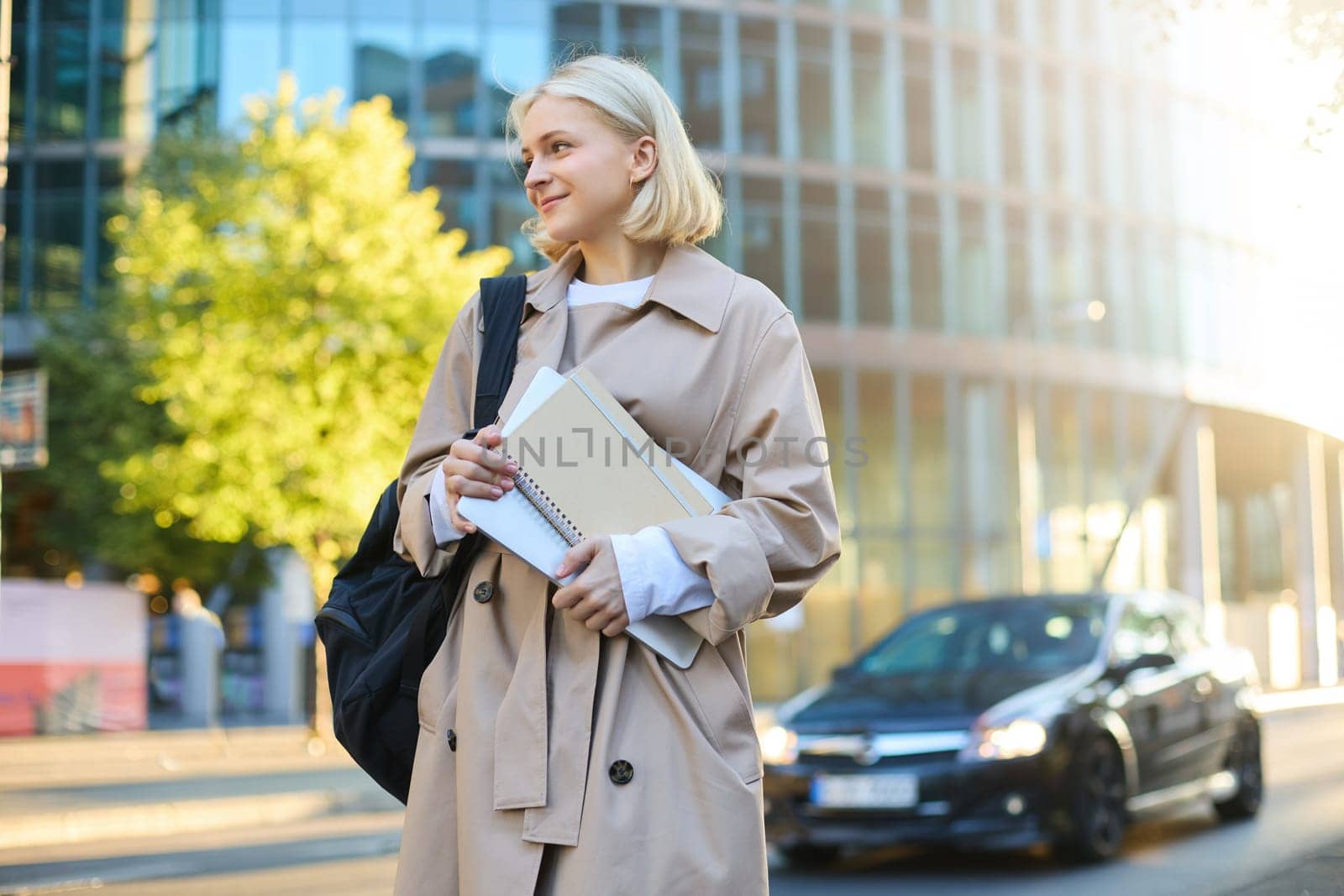 College lifestyle and student concept. Smiling blonde woman on street, sunny day, skyscraper behind her, girl holding documents, notebooks and backpack.