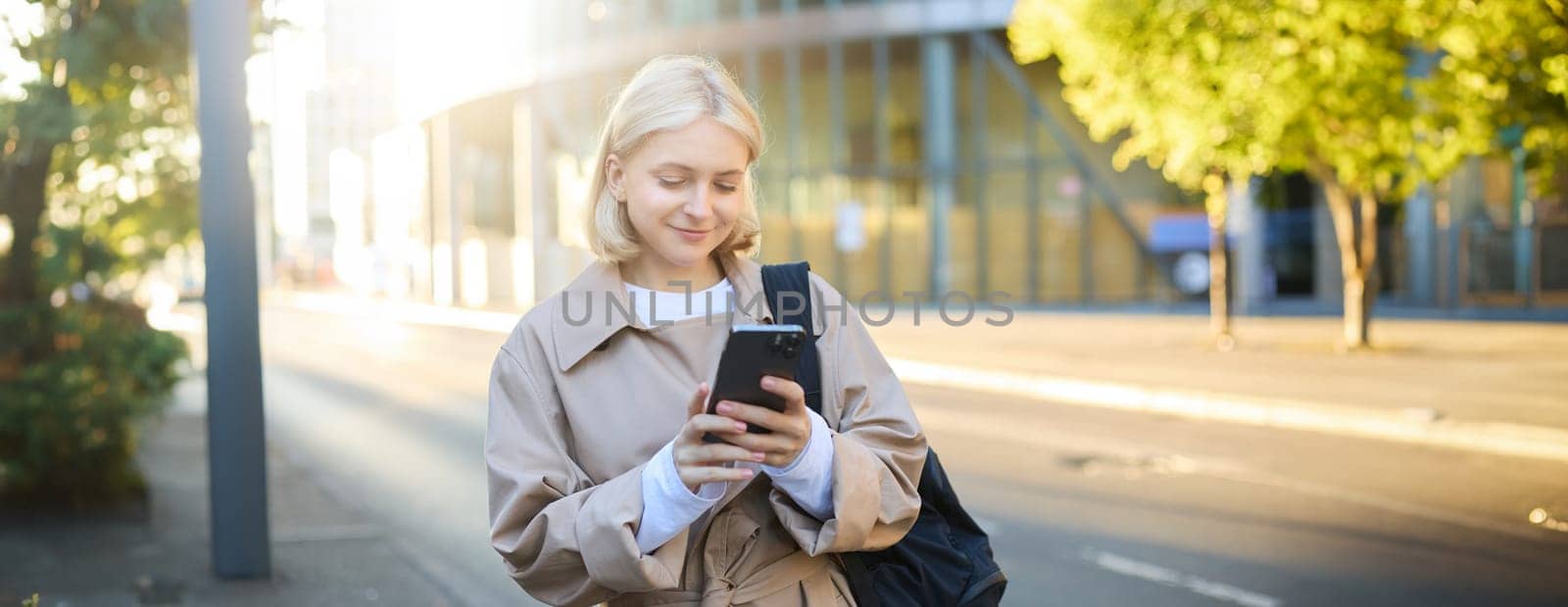 Street style portrait of blonde smiling woman walks on street, sunny bright day, holding smartphone, carries backpack, orders taxi on mobile application, sends message on cell phone.