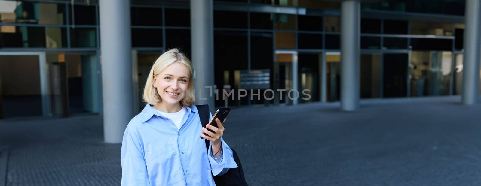 University and people concept. Vertical portrait of young woman standing near office building on street, student in college campus, holding smartphone, laptop and backpack on shoulder by Benzoix