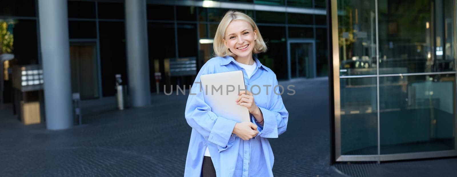 Image of young female employee, working woman standing near her office on street, holding pile of documents and smiling. People and lifestyle concept