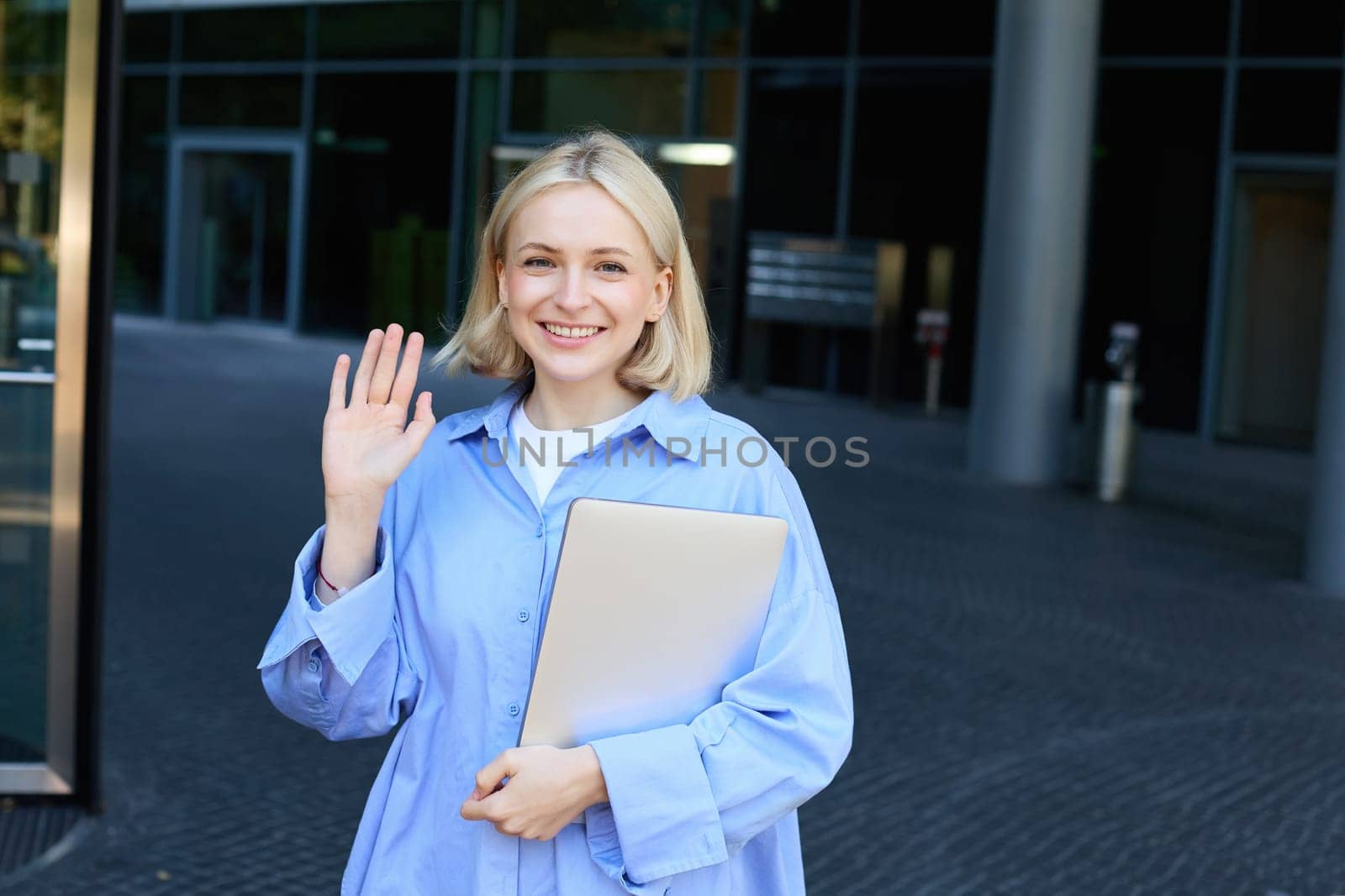 Friendly smiling student, blond girl with notebooks, tutor waving hand at camera, saying hello, standing near office building outdoors by Benzoix