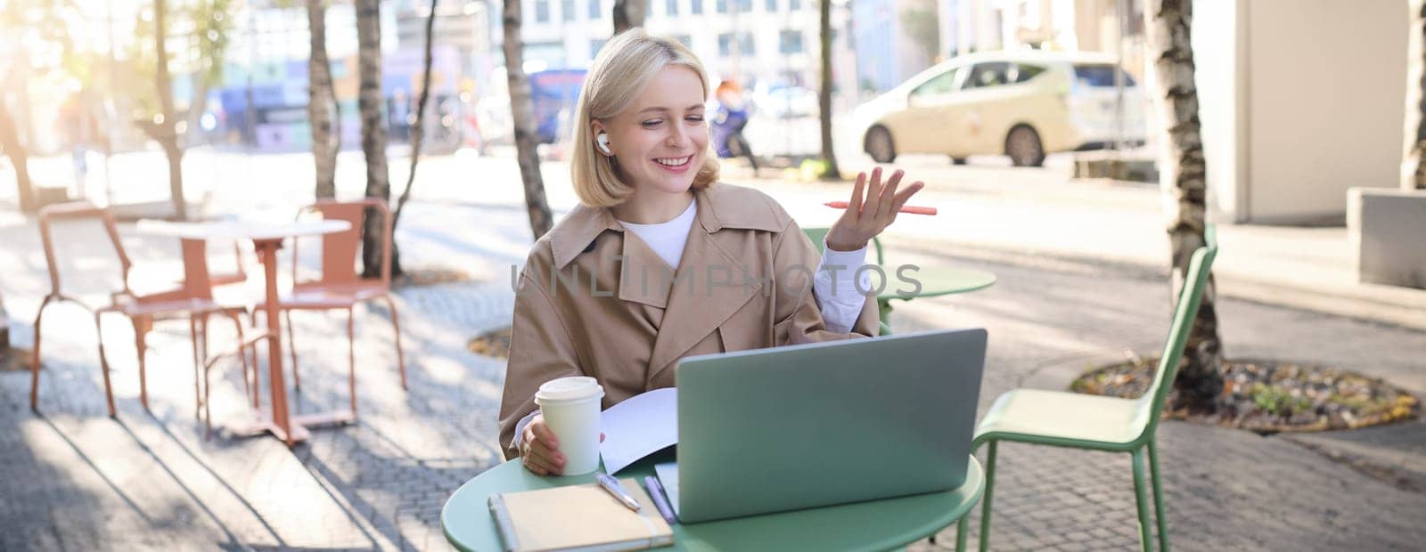 Friendly, smiling blond woman chatting online, wearing wireless headphones, looking at laptop and gesturing, talking to someone, working remotely by Benzoix