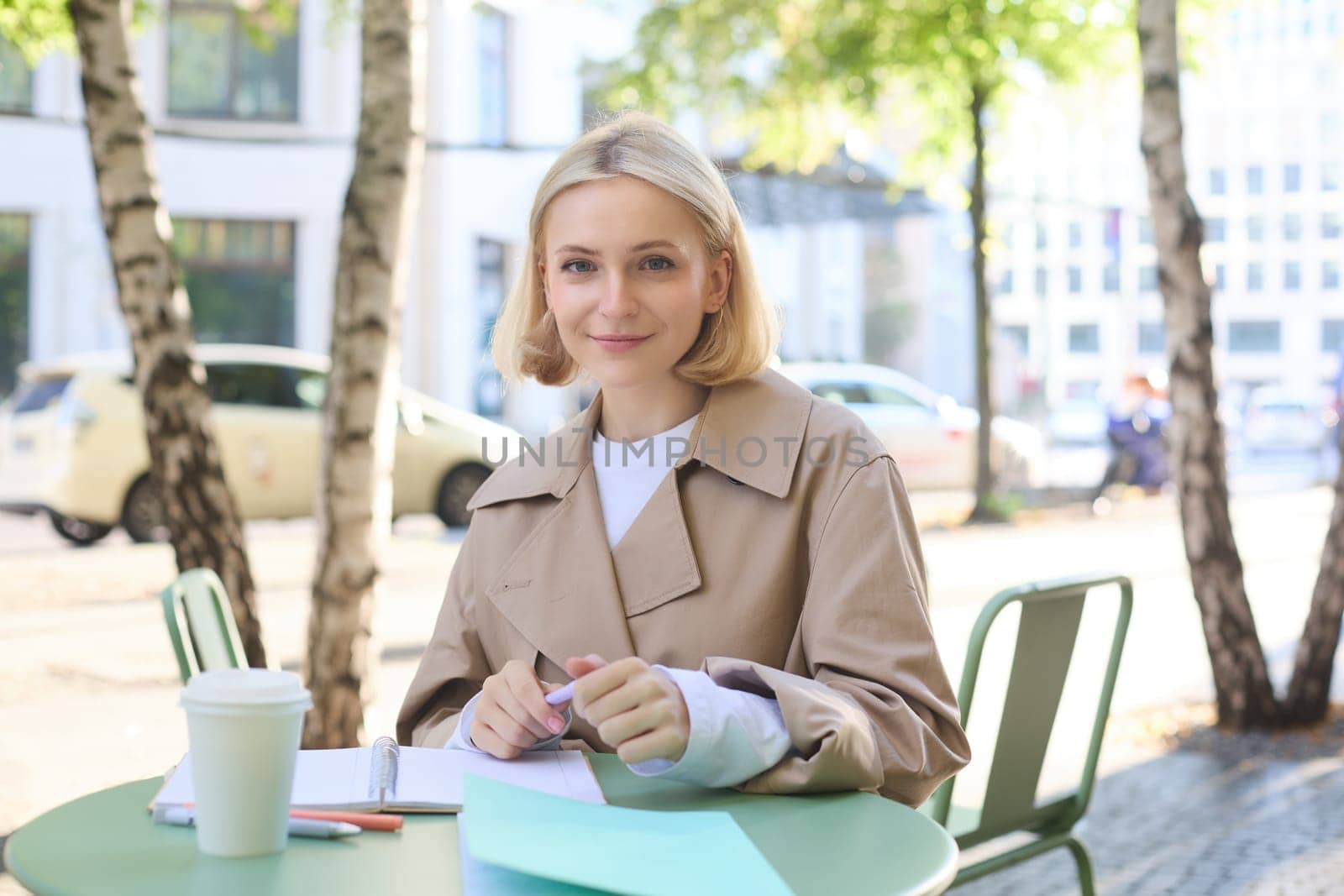 Portrait of young stylish student, woman working outdoors in cafe, doing homework, writing essay, holding markers, smiling at camera.