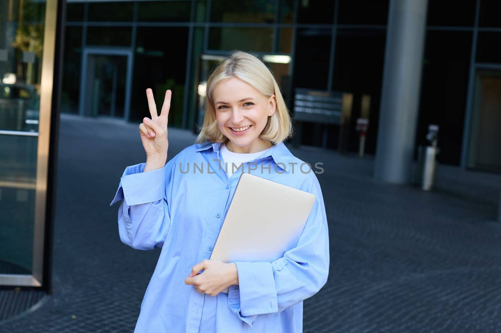 Young positive female model, student with notebooks and study material showing peace sign, standing near university, college campus, promo for new education scheme.