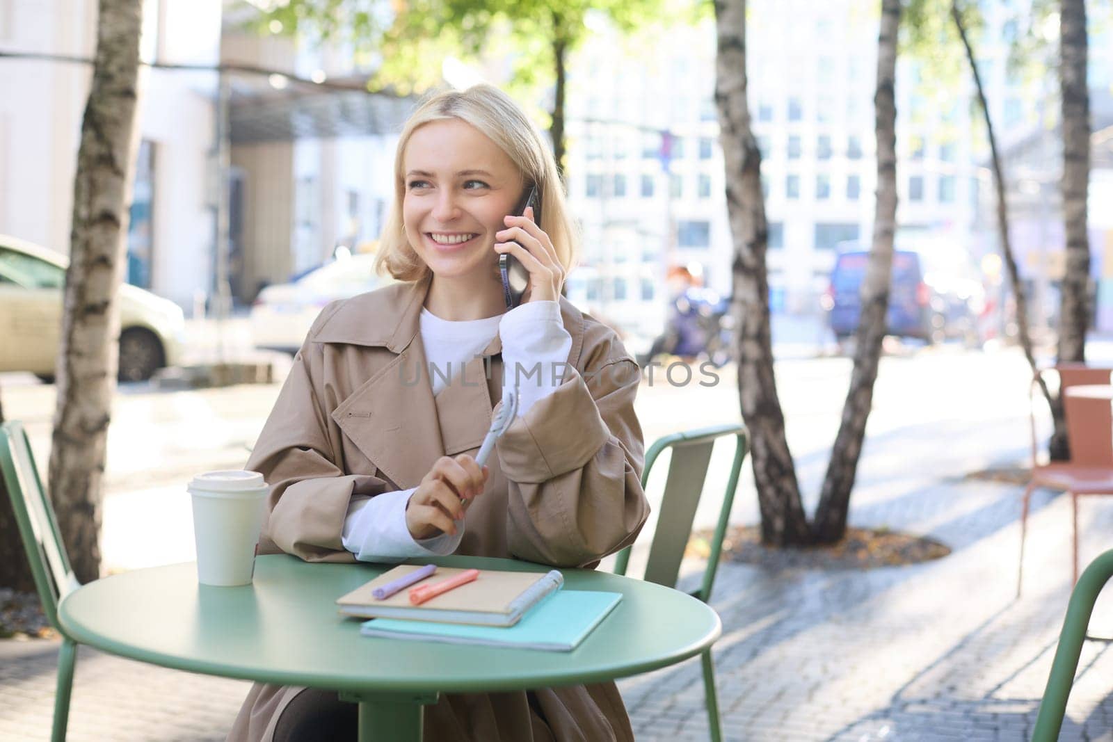 Image of young modern female model in trench, sitting in cafe outdoors, has journals and notebooks on table, talking on mobile phone, answers a call and smiles.