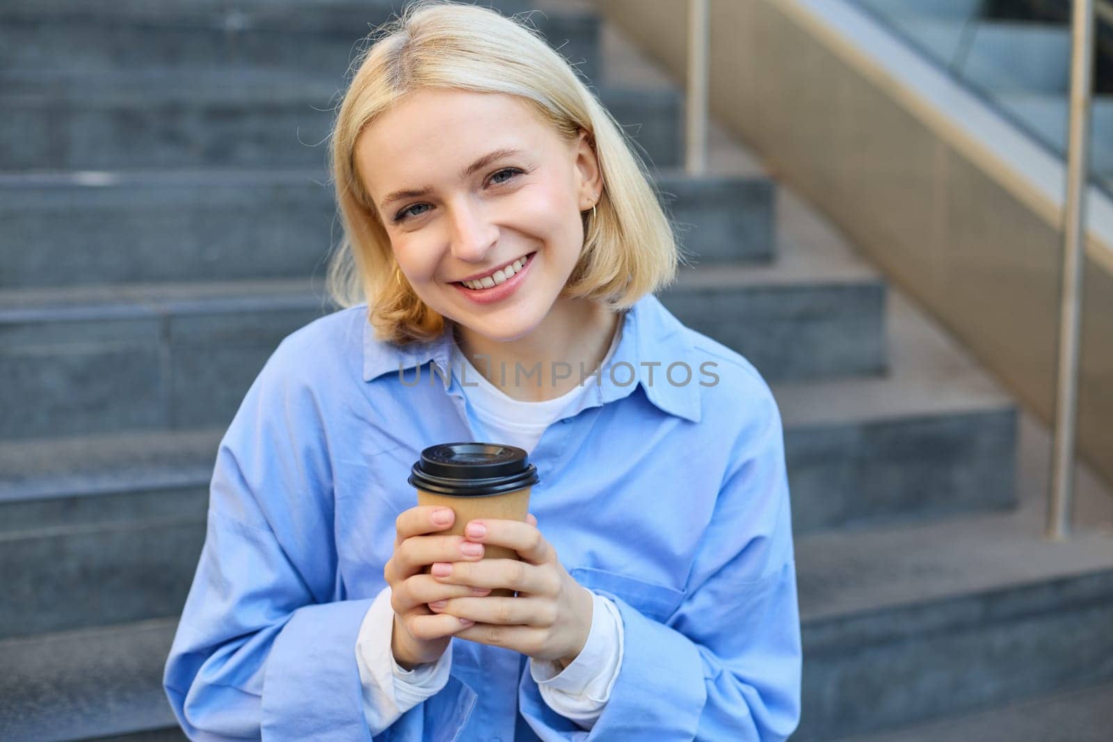 Close up portrait of female model, cute student with cup of coffee, sitting on stairs near campus, spending time outside.