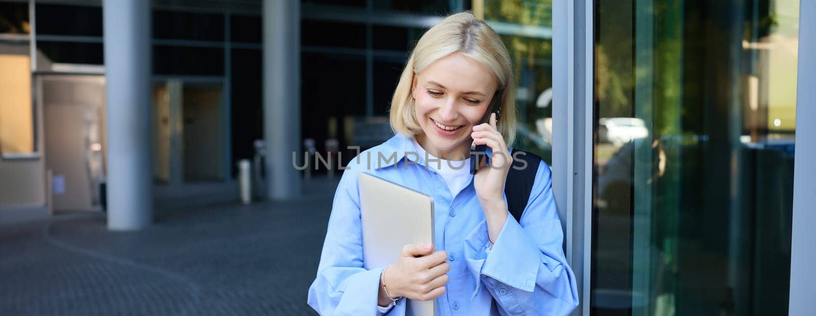 Close up portrait of smiling blond woman, holding laptop, has backpack on shoulder, talking on smartphone, answer a phone call by Benzoix