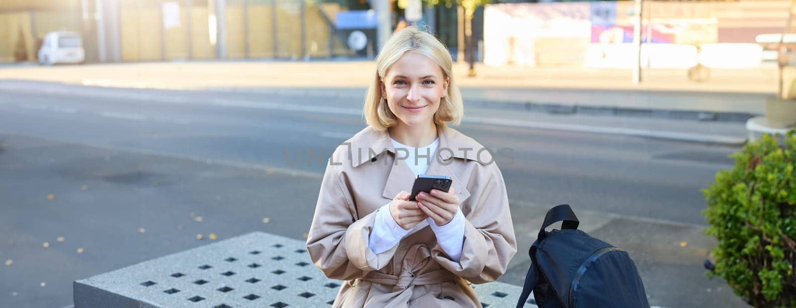 Outdoor shot of young blonde woman on street, sitting on bench with mobile phone, smiling at camera, waiting for friend outside of building, having a break by Benzoix