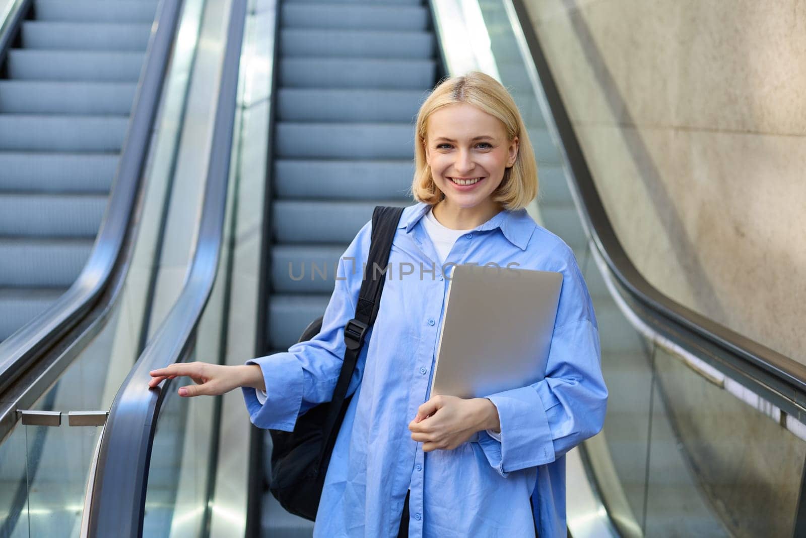 Street style portrait of woman on escalator, holding laptop and backpack on her shoulder, smiling at camera, walking in city by Benzoix