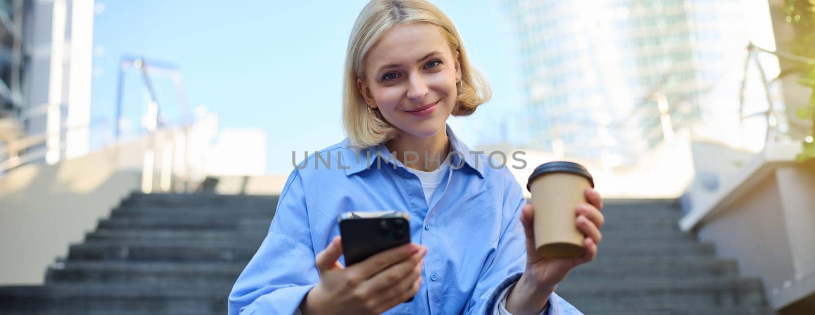 Portrait of beautiful blond woman, smiling girl student drinking coffee, sitting on stairs with smartphone, looking happy at camera.