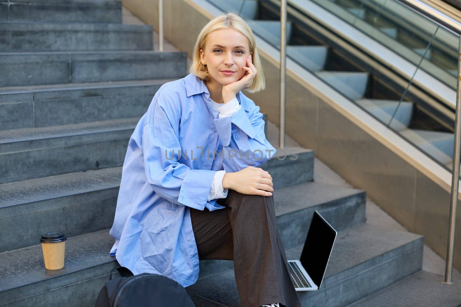 Portrait of young modern woman, student or freelancer, sitting on outdoor stairs, resting in city, has laptop and coffee, looking at camera with confident, smiling face.
