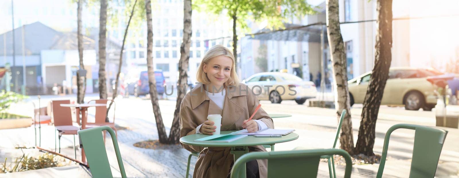 Smiling, cheerful woman drinking her coffee in street cafe, writing in notebook, doing homework, making notes. Student and lifestyle concept by Benzoix