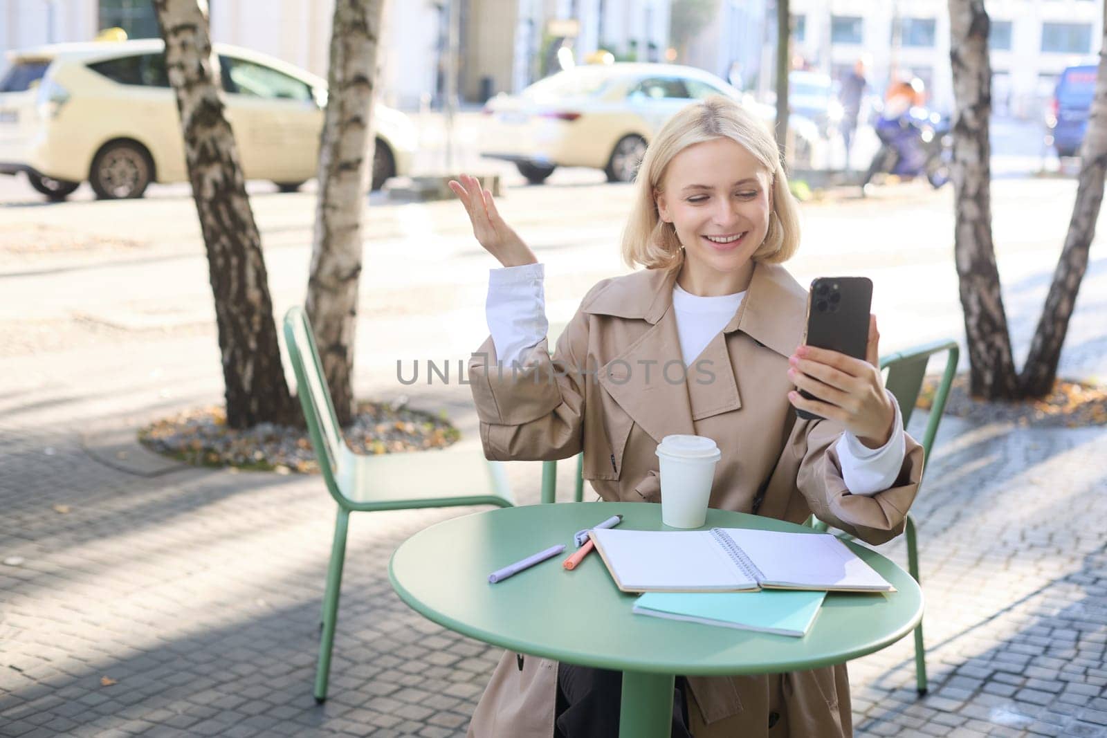 Portrait of young stylish woman in trench, sitting outdoors in city cafe, talking on video chat, using mobile phone to connect to online conversation, drinking coffee by Benzoix