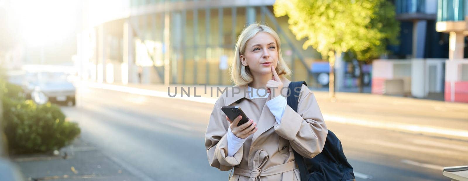 Portrait of woman with thinking face, standing with backpack on street, holding smartphone, pondering, making a decision with thoughtful face by Benzoix