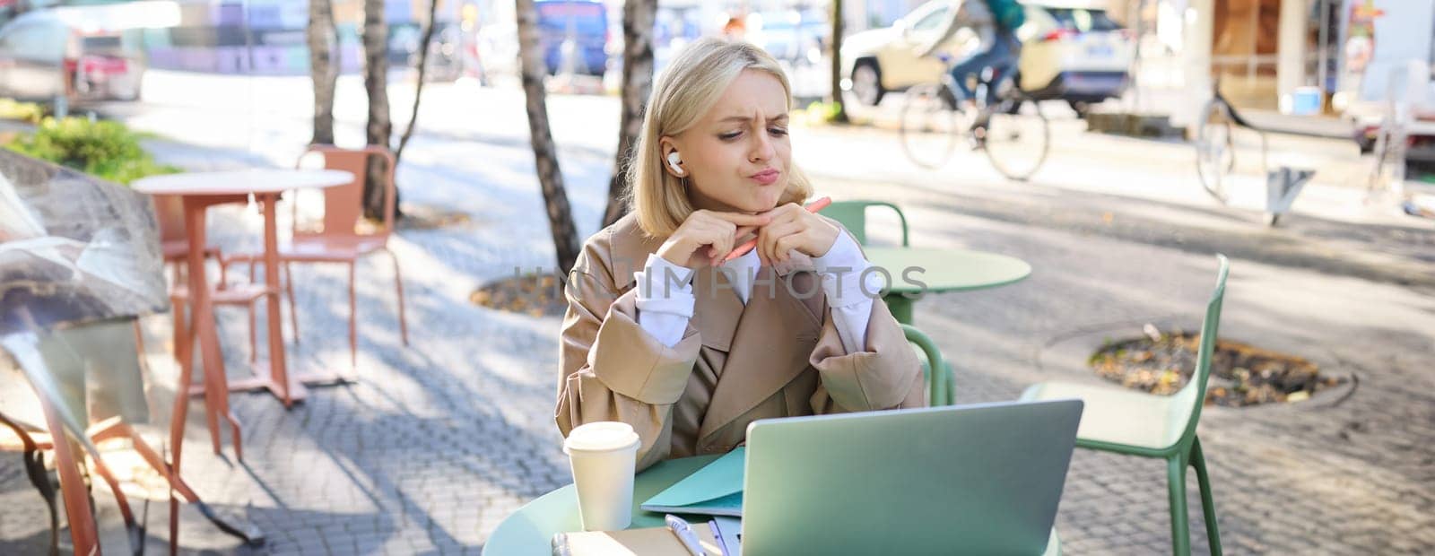 Portrait of working woman in outdoor cafe, wearing wireless headphones, student frowning while looking at laptop, confused by lesson theme.