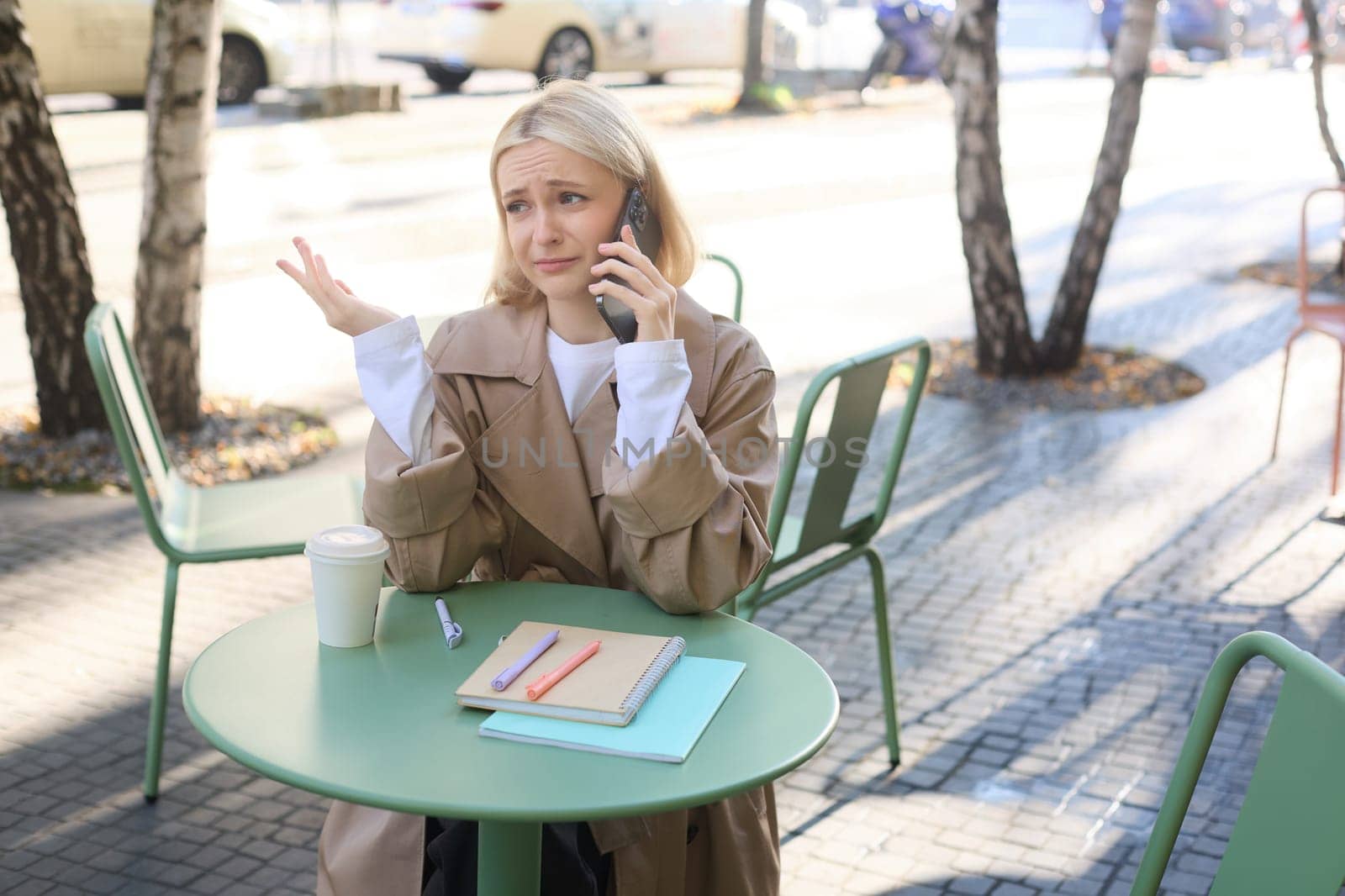 Image of young upset woman in cafe, talking on mobile phone with sad, disappointed face expression, raising hand up unhappy.