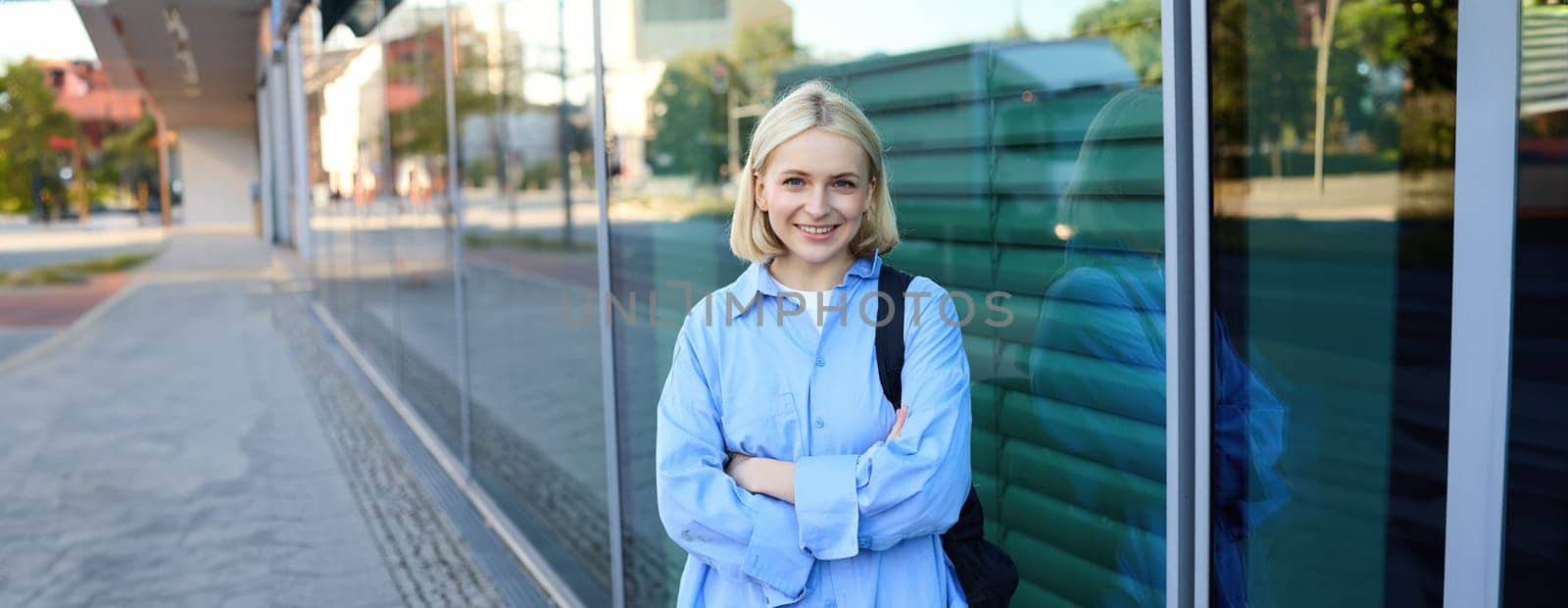 Image of young modern woman on street, college student in blue shirt, with backpack, cross arms on chest and smiles with confidence at camera.