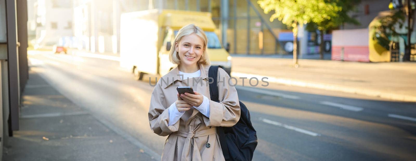 Street style portrait of blonde smiling woman walks on street, sunny bright day, holding smartphone, carries backpack, orders taxi on mobile application, sends message on cell phone.