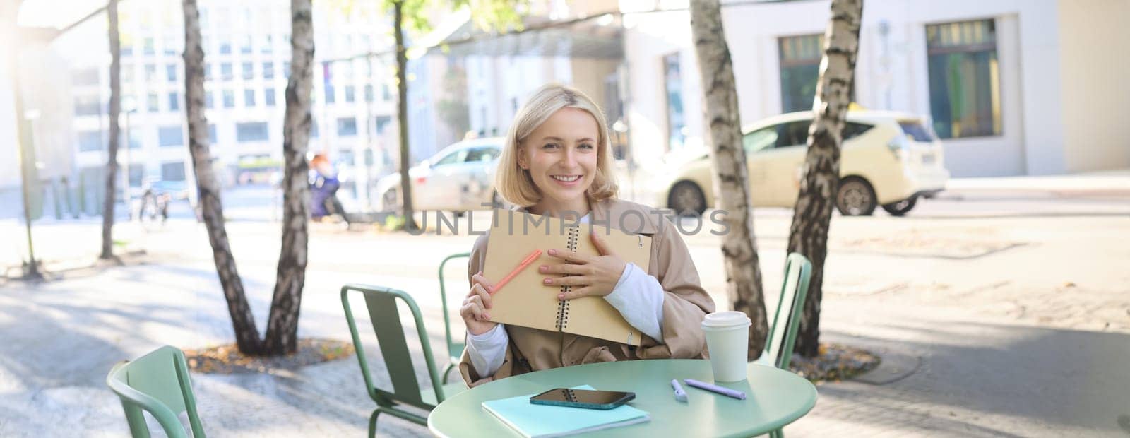 Image of young modern woman, student sitting in coffee shop with smartphone, holding notebook, showing her yellow journal, writing in planner and smiling by Benzoix