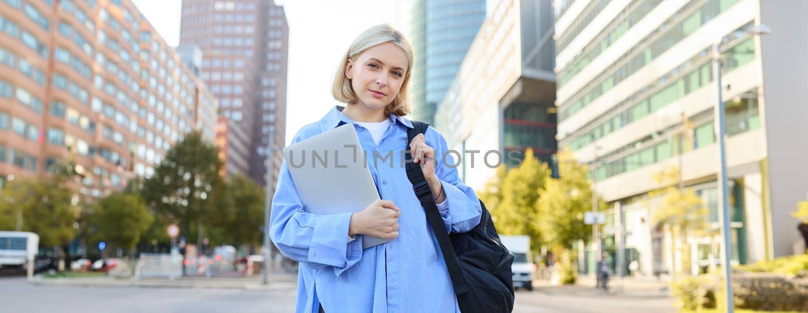 Portrait of young confident woman, college student with backpack and laptop, heading to lesson, standing outdoors on empty street with big buildings behind by Benzoix