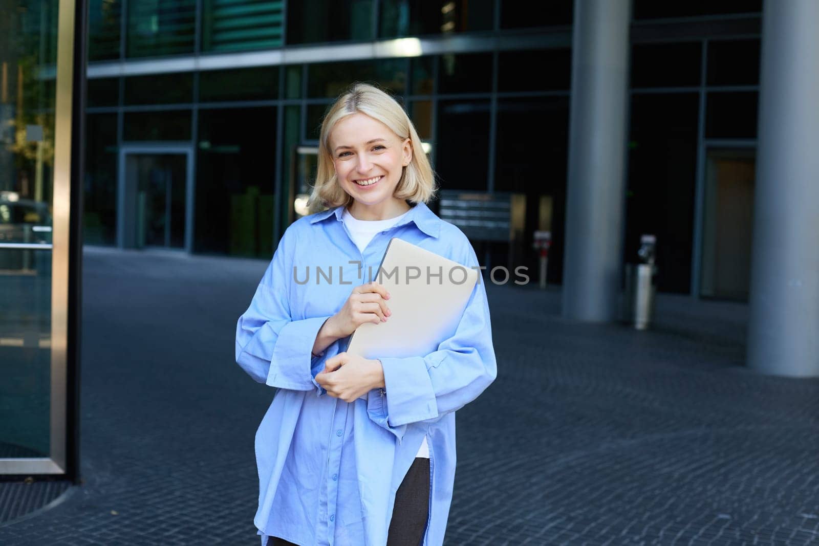 Portrait of young blond woman, student standing near her campus with notebooks and documents, wearing blue shirt and smiling at camera by Benzoix