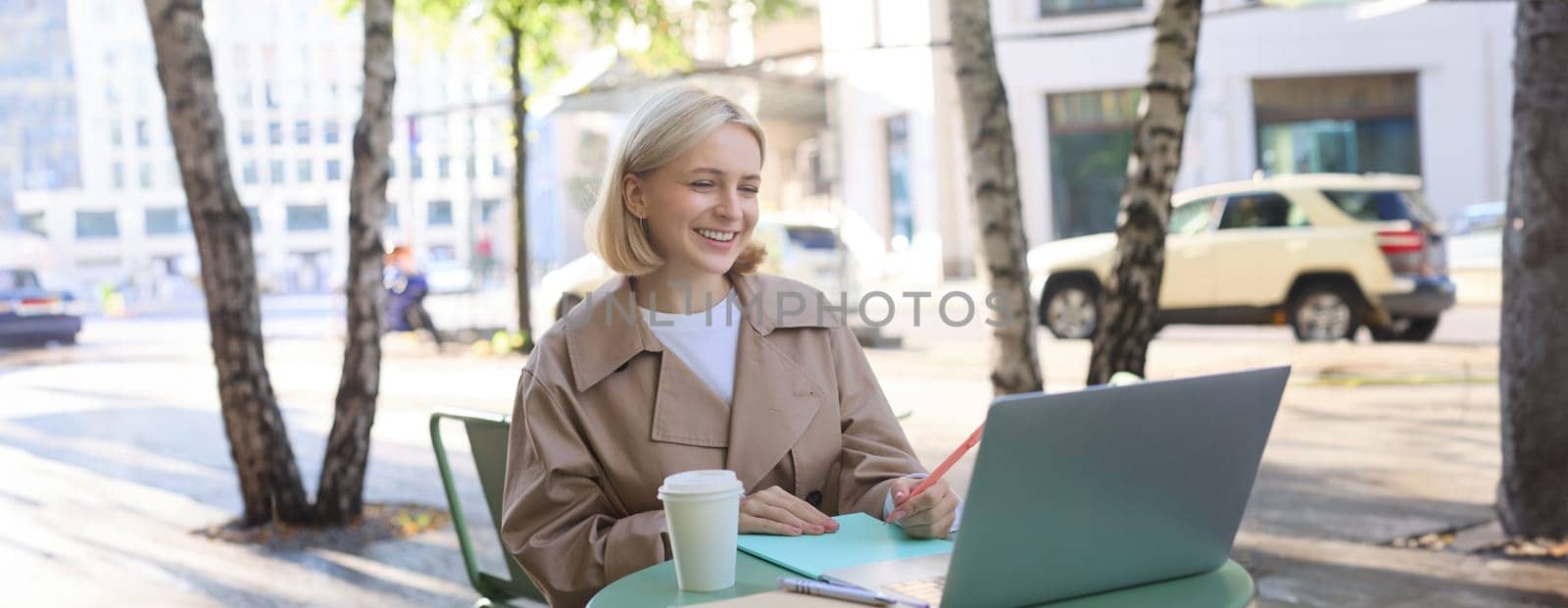 Remote workplace. Smiling young woman freelancer, student using laptop in outdoor cafe, talking to someone, attends online lecture, drinking coffee by Benzoix