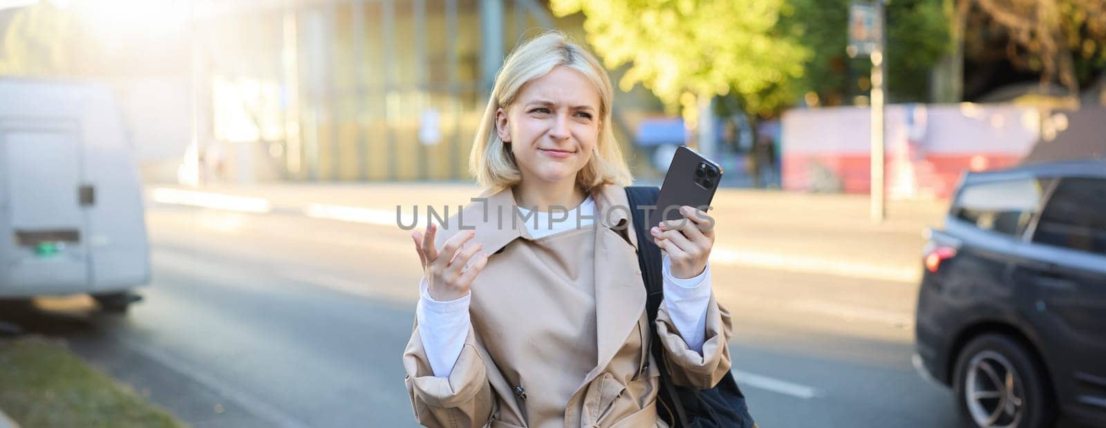 Portrait of confused blonde woman on street, carries backpack, holding smartphone, frowning and looking puzzled, unsure where to go, using mobile map by Benzoix