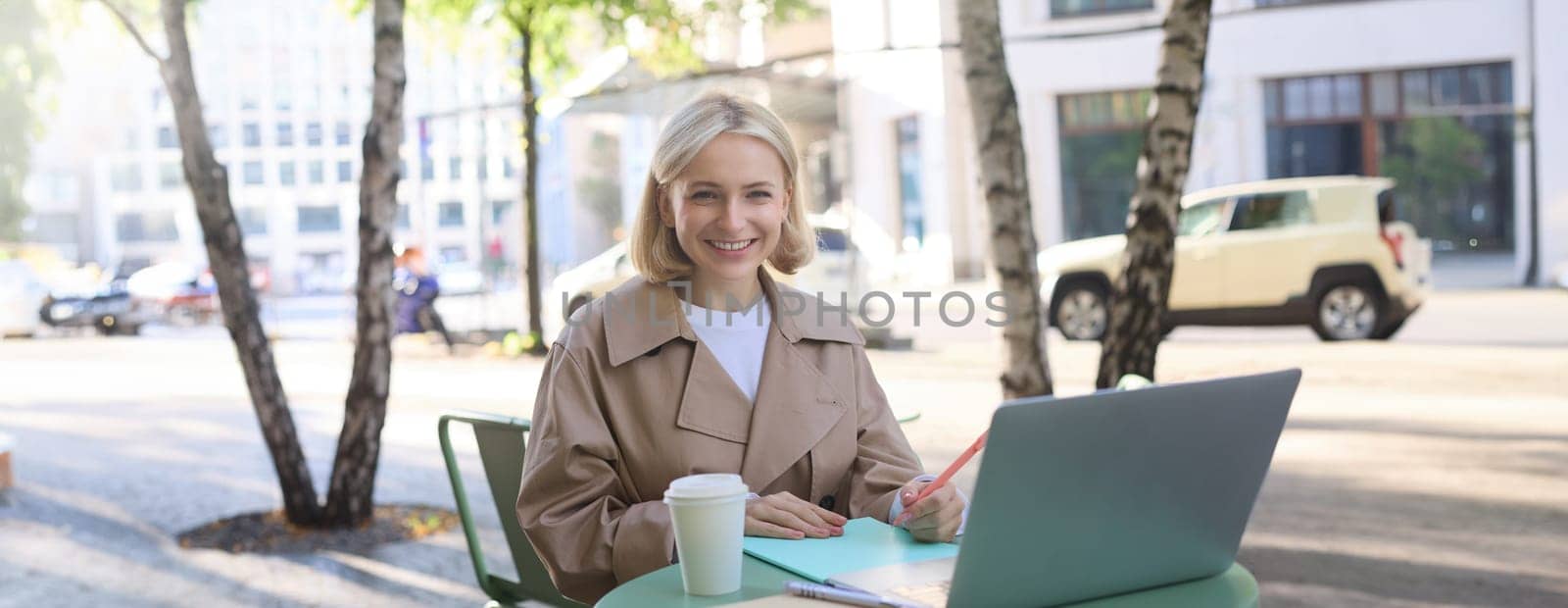Image of young stylish woman, sitting on street, drinking coffee in cafe, working remotely on laptop, attends online course or doing homework by Benzoix