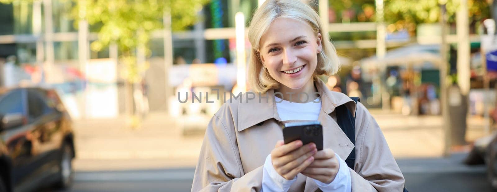 Close up portrait of young smiling blond woman, student on street, using mobile phone, checking messages on smartphone, walking on road by Benzoix