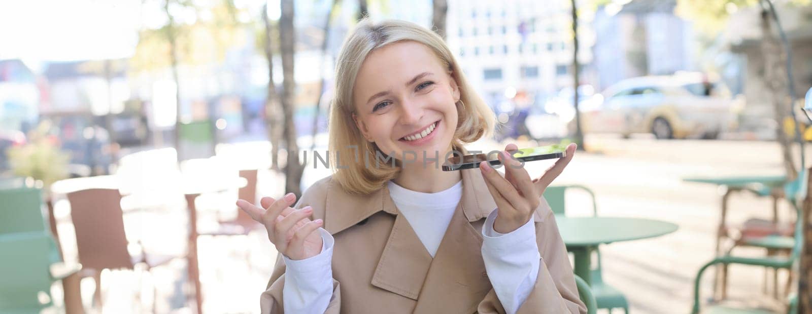 Close up portrait of smiling blond woman, girl with mobile phone outdoors, sitting in cafe, recording voice message, talking into smartphone with microphone near mouth.