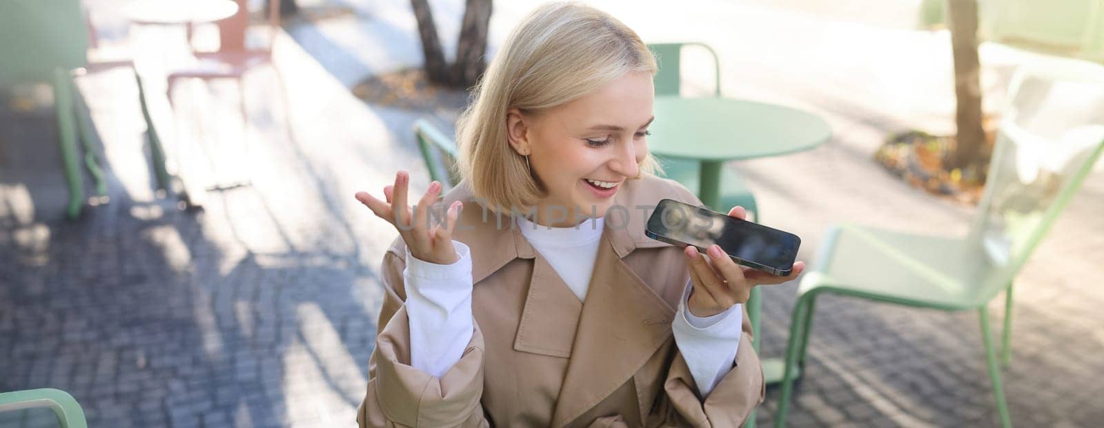 Lifestyle portrait of happy young woman, sitting in cafe and recording voice message, holding mobile phone near lips and talking into microphone, spending time in cafe, drinking coffee and chatting.