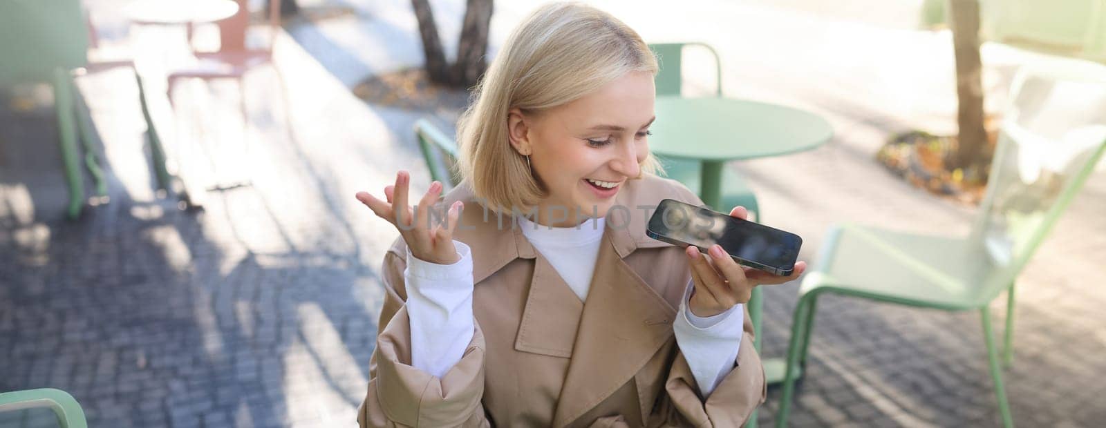 Lifestyle portrait of happy young woman, sitting in cafe and recording voice message, holding mobile phone near lips and talking into microphone, spending time in cafe, drinking coffee and chatting by Benzoix