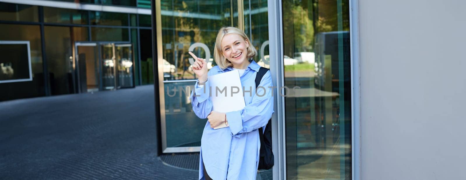 Portrait of smiling young woman, student or office worker with laptop, pointing finger at upper right corner, showing building and looking happy.