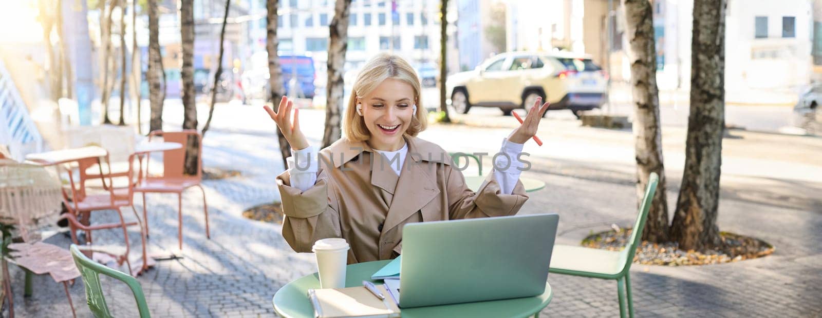 Enthusiastic young woman hear great news during online meeting, looking at laptop and celebrating, raising hands up to cheer, sitting in outdoor cafe.