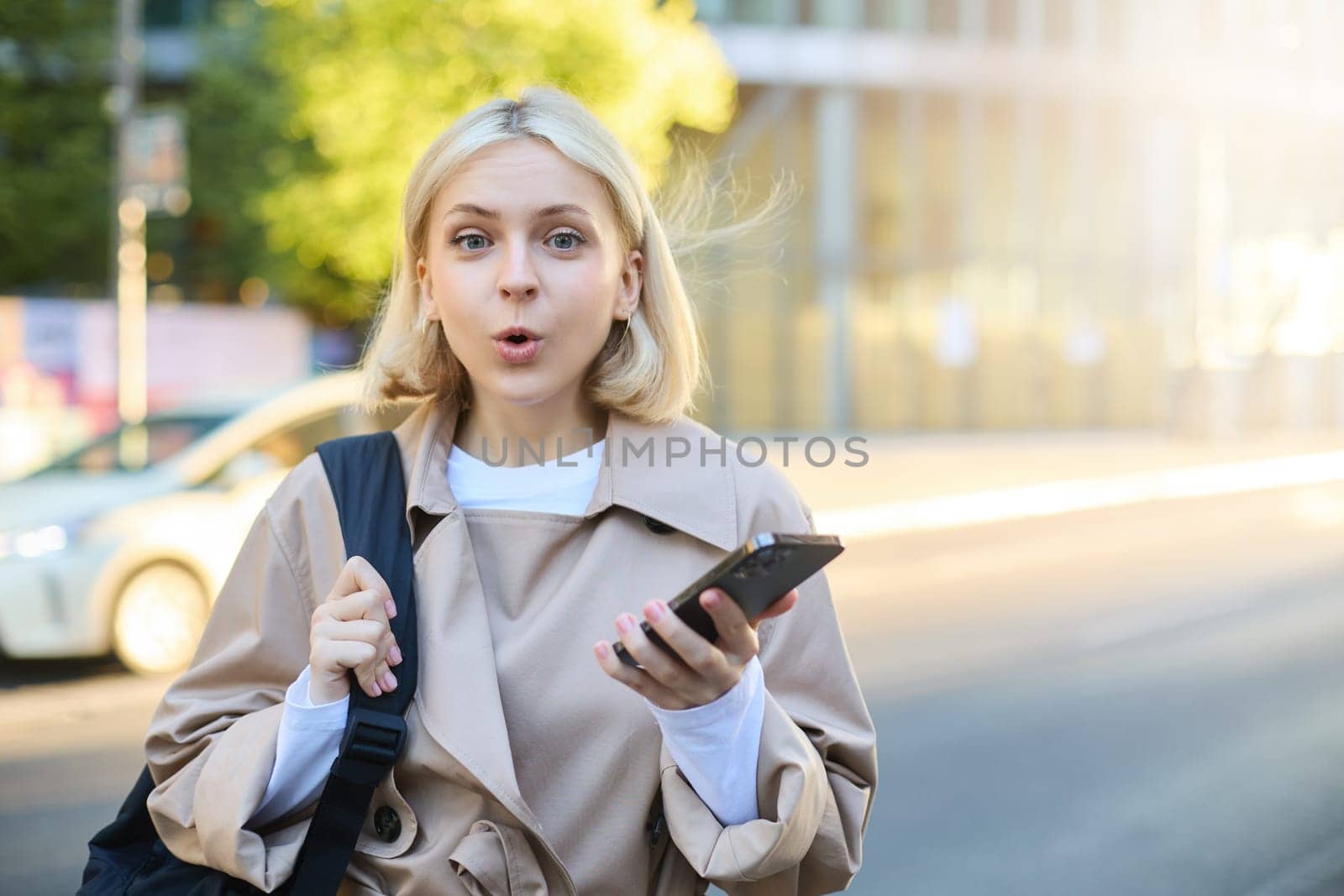 Image of young surprised woman, standing on street with mobile phone, looking amazed at camera, saying wow.