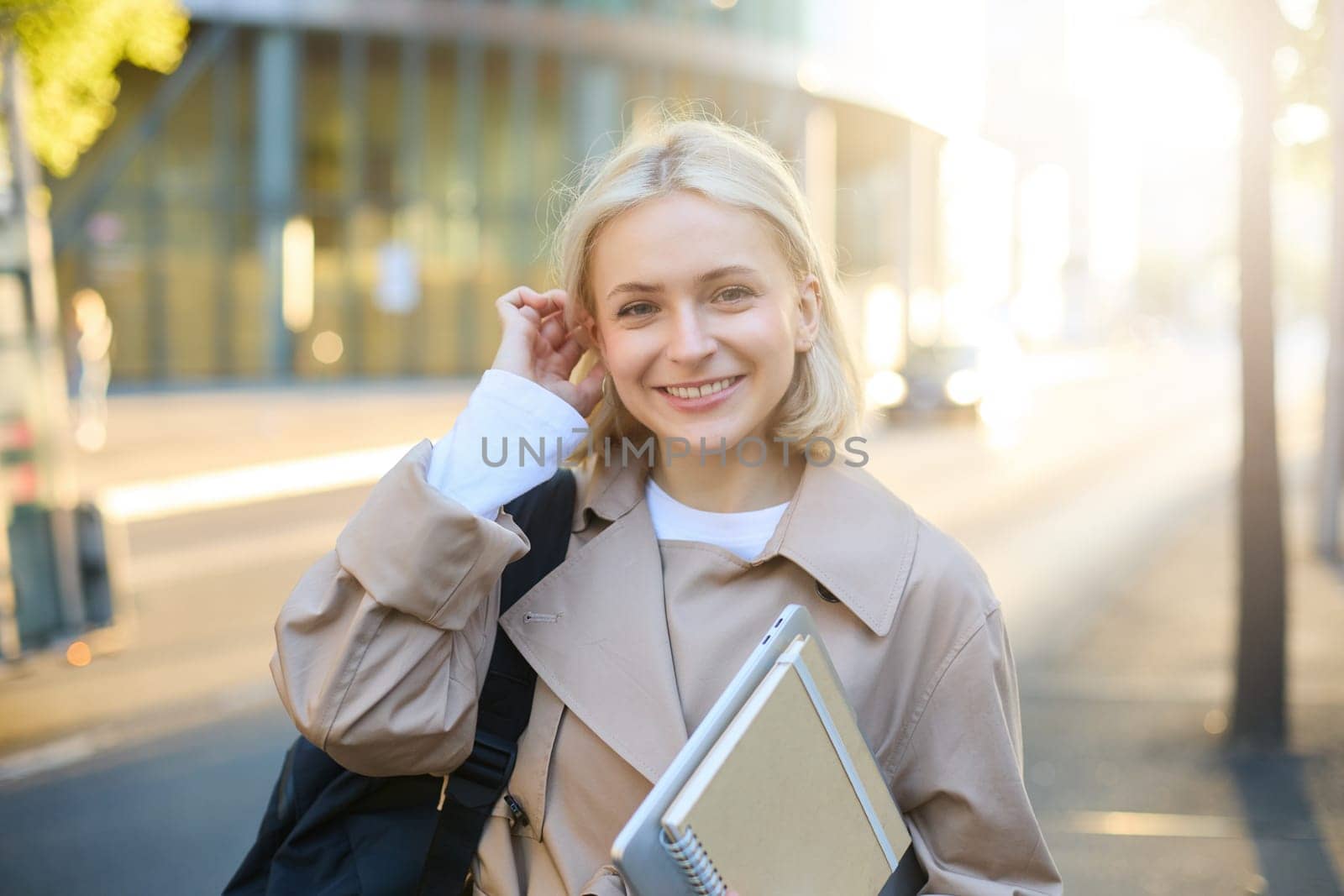 Young blonde woman, college student on street, holding journals and notebook, carry her homework material, going to university, smiling and looking happy by Benzoix