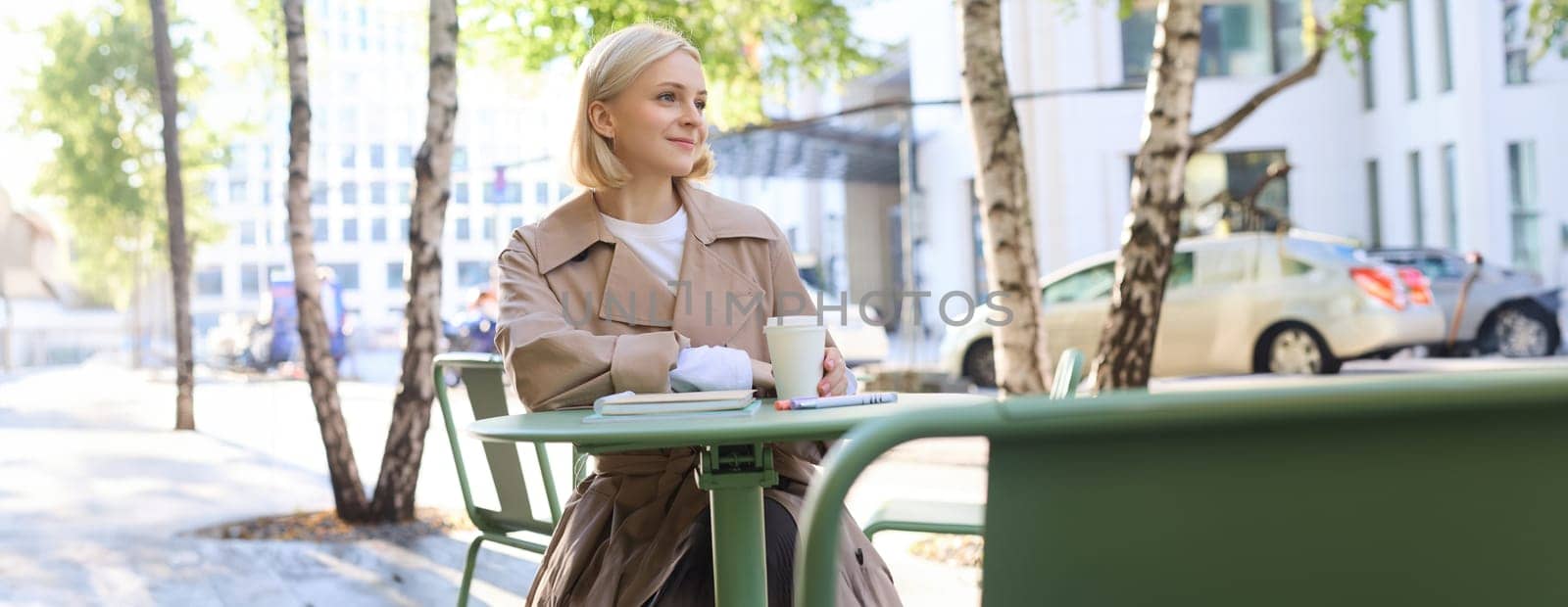 Urban lifestyle concept. Young beautiful woman sitting in outdoor cafe, contemplating city life, drinking coffee and smiling on street.