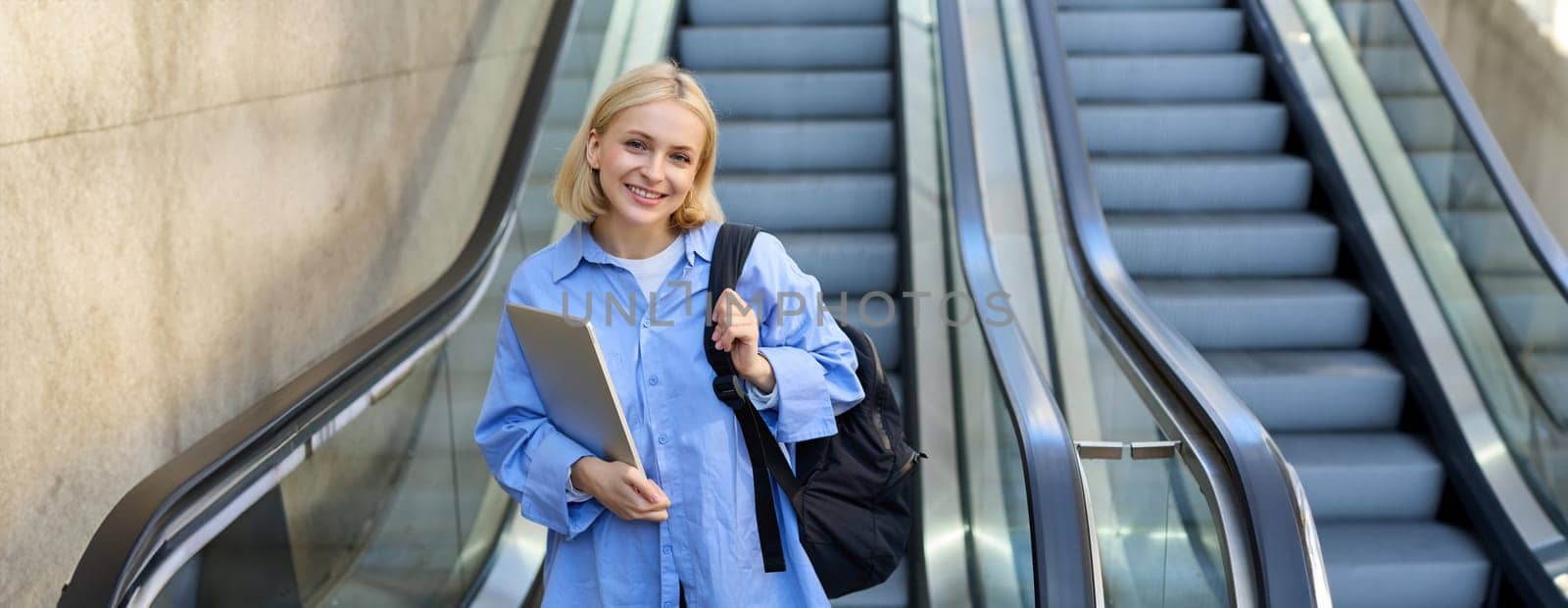 Image of young modern woman, student with backpack, carries her laptop in hands, stands near escalator, walks in city.