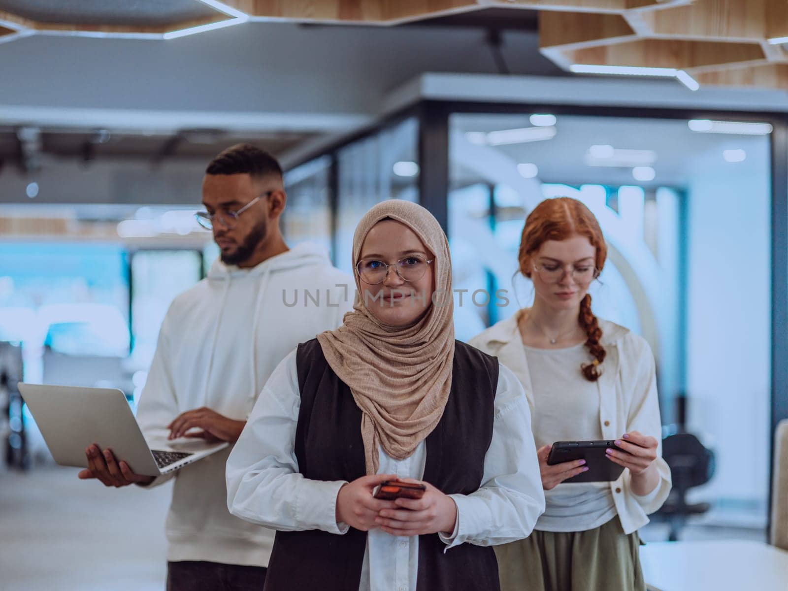 A diverse group of young businessmen, including a Muslim woman wearing a hijab, an orange-haired woman, and an African American, stand together in a modern office, showcasing a vibrant and inclusive workspace by dotshock