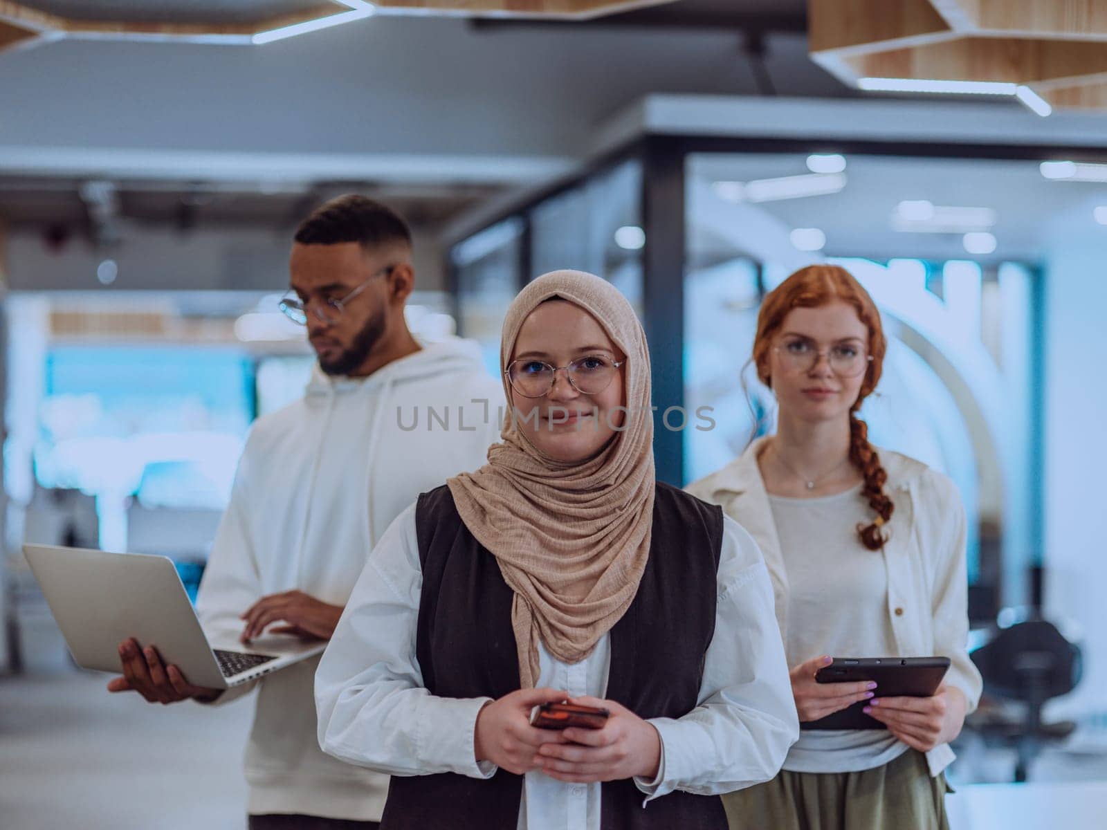 A diverse group of young businessmen, including a Muslim woman wearing a hijab, an orange-haired woman, and an African American, stand together in a modern office, showcasing a vibrant and inclusive workspace by dotshock