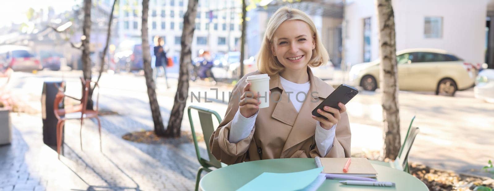 Portrait of blond smiling woman sitting in cafe, enjoying cup of takeaway coffee, using mobile phone, working on smartphone, social media blogger making post online.