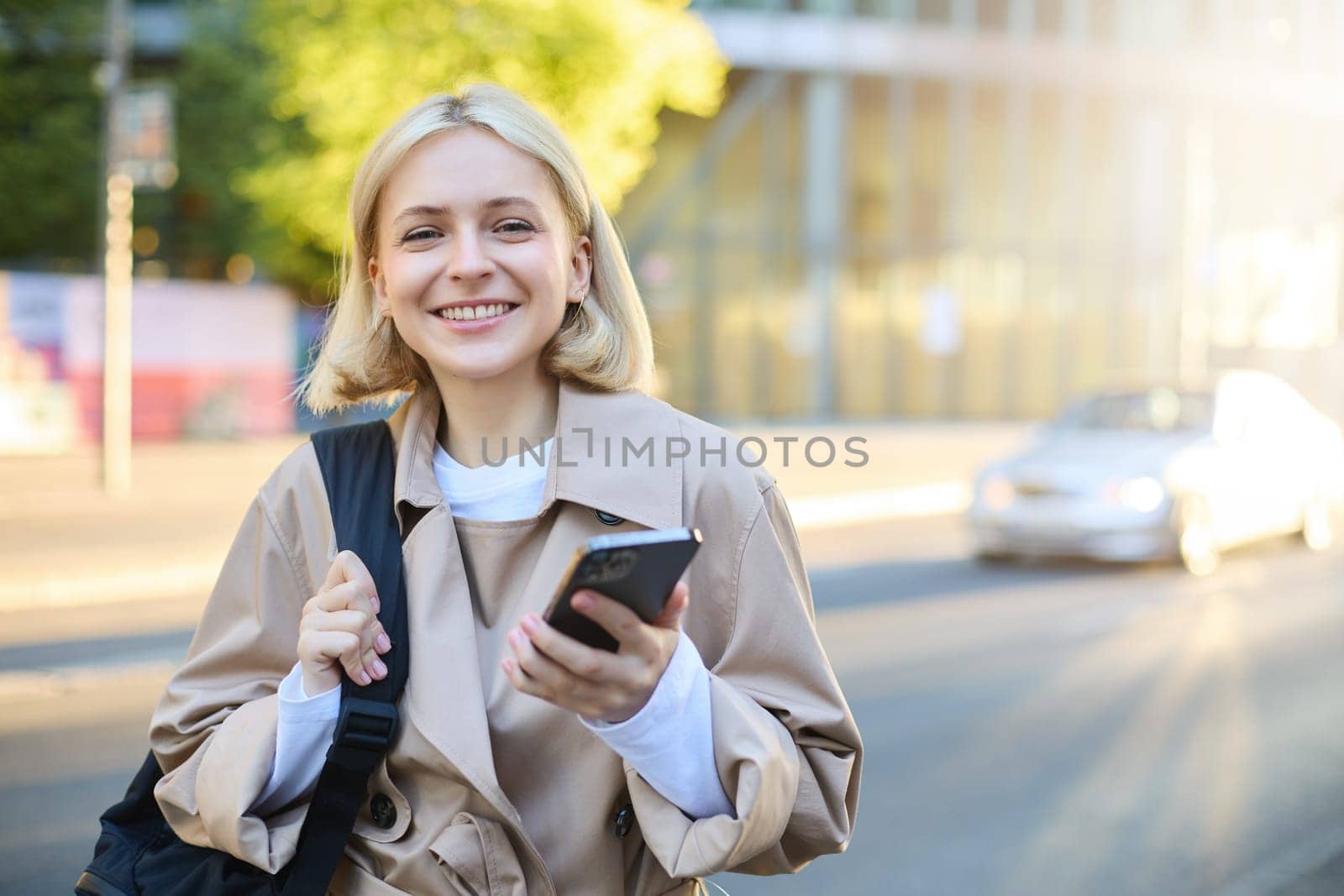 Close up portrait of happy, beautiful young woman with backpack, standing on street and using mobile phone, waiting for taxi near road, smiling.