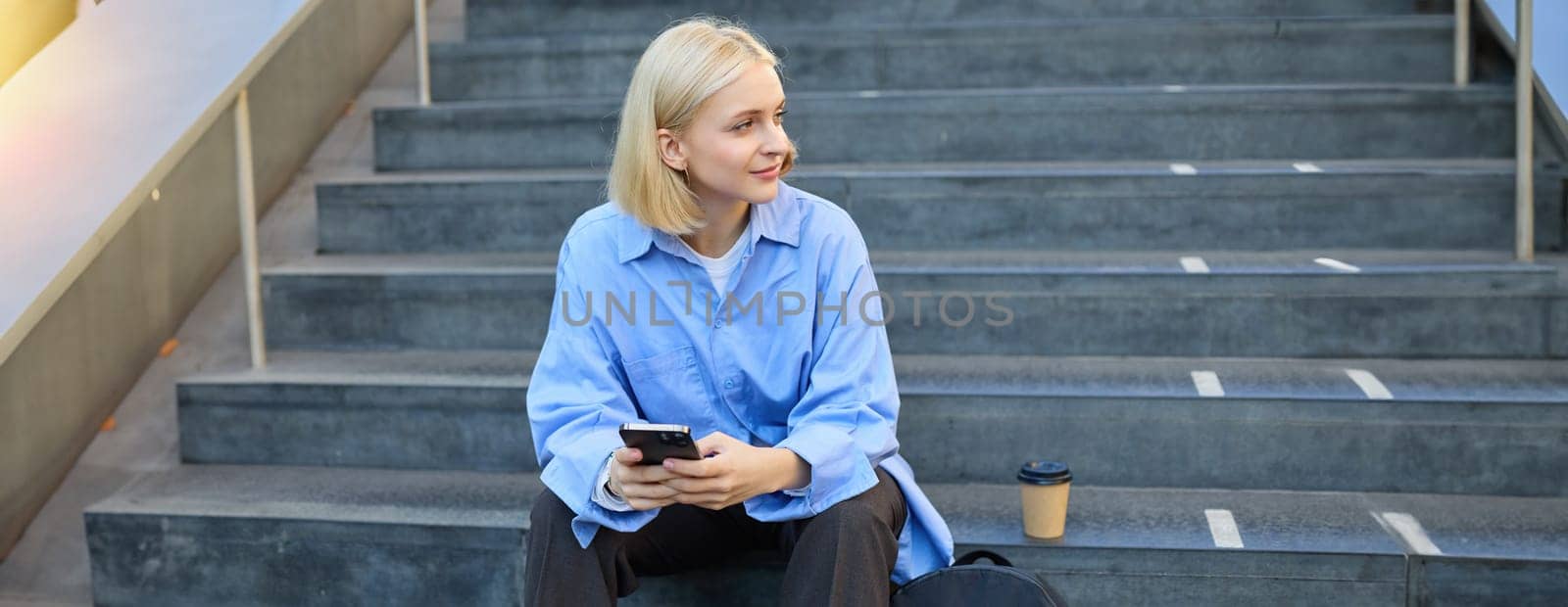 Lifestyle portrait of young urban female model, student drinks cup of takeaway coffee, sitting on stairs outdoors, using mobile phone, smiling and looking happy while taking a break by Benzoix