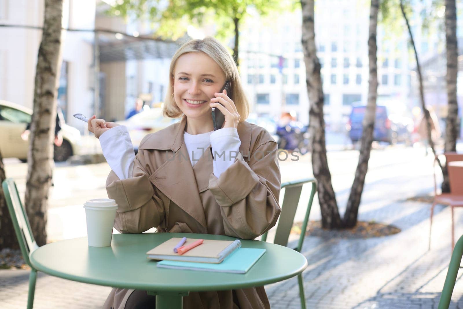 Image of stylish blond woman talking on mobile phone, drinking coffee in cafe outdoors, enjoying warm weather in city centre, answer telephone call on smartphone by Benzoix