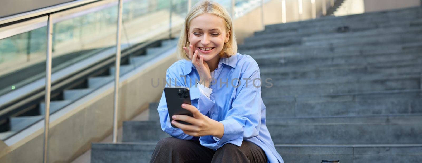 Image of young happy woman, sitting on stairs outside on street, taking selfie on smartphone camera, posing for photo social media profile, smiling.