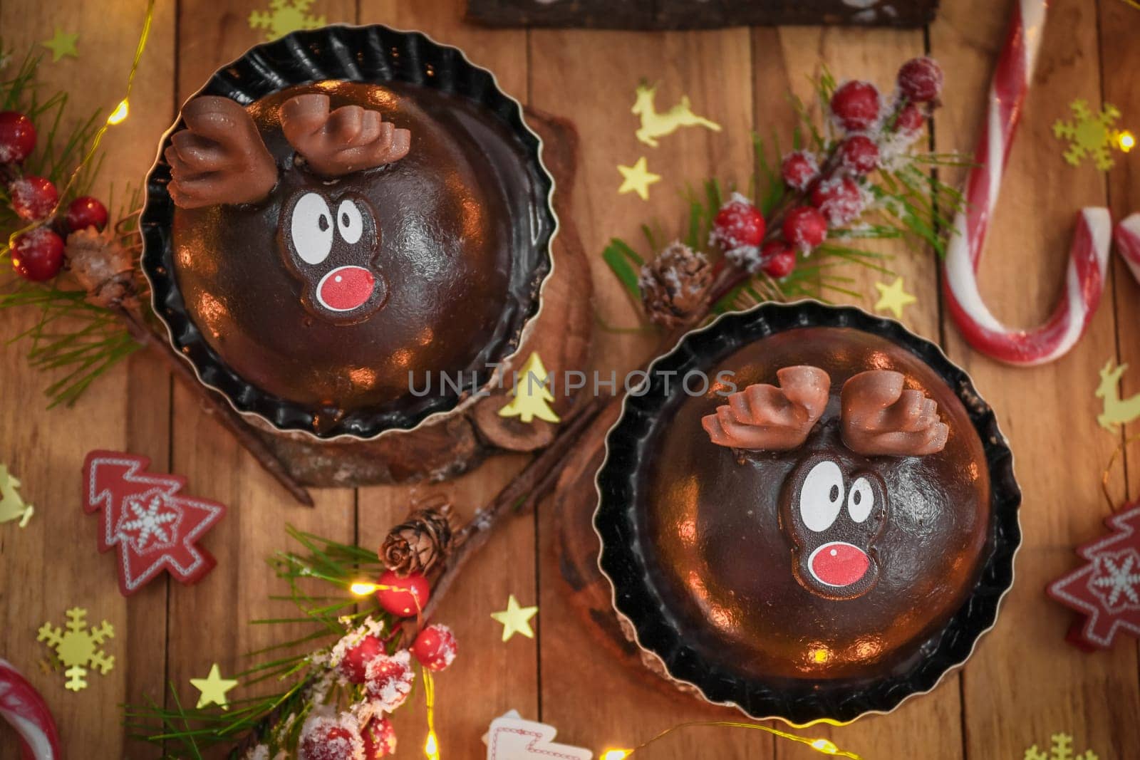 Two chocolate deer in a wooden tray with sweets, confetti, a burning garland and a christmas decoration on a brown wooden table, close-up top view.